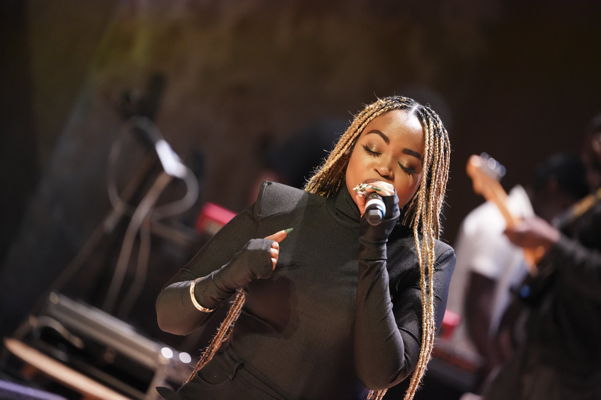 Lenaa I AM performing live on stage, singing into a handheld microphone with her band behind her